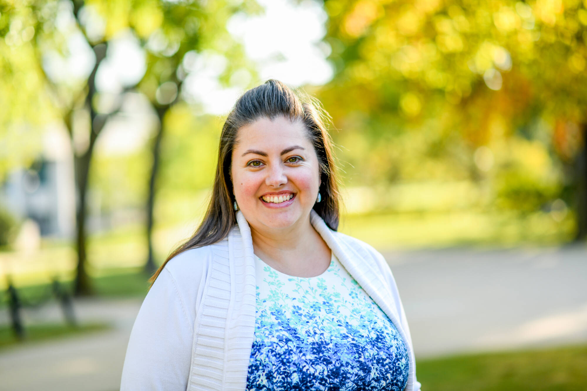 Picture of Samantha Mayse. Samantha has brown hair, is wearing a blue shirt with a white sweater, and is posed in front of trees with lots of sunshine.